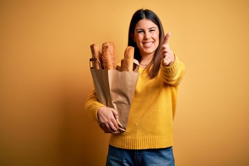 Young beautiful woman holding a bag of fresh healthy bread over yellow background smiling friendly offering handshake as greeting and welcoming. Successful business.