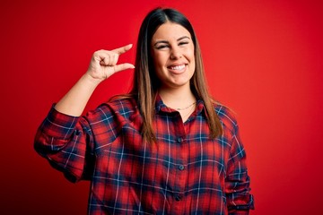 Young beautiful woman wearing casual shirt over red background smiling and confident gesturing with hand doing small size sign with fingers looking and the camera. Measure concept.