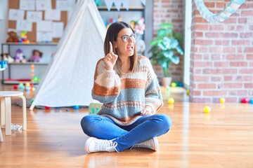 Young beautiful teacher woman wearing sweater and glasses sitting on the floor at kindergarten...