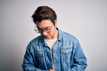 Young handsome chinese man wearing denim jacket and glasses over white background with hand on stomach because indigestion, painful illness feeling unwell. Ache concept.