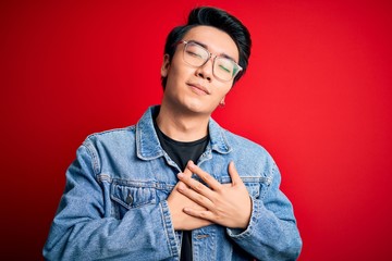 Fototapeta na wymiar Young handsome chinese man wearing denim jacket and glasses over red background smiling with hands on chest with closed eyes and grateful gesture on face. Health concept.