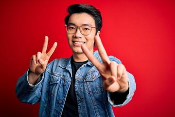Young handsome chinese man wearing denim jacket and glasses over red background smiling looking to...