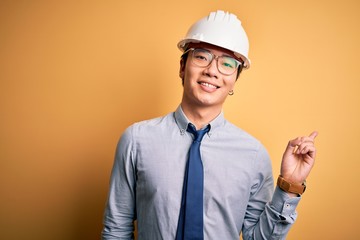 Young handsome chinese architect man wearing safety helmet and tie over yellow background with a big smile on face, pointing with hand and finger to the side looking at the camera.