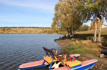 There is an old catamaran on the Bank of the autumn pond. There are a number of trees along the...