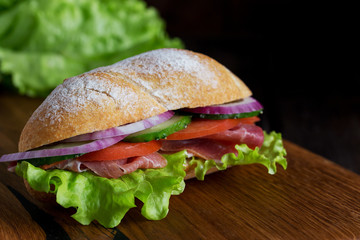 Fresh an tasty submarine sandwich with prosciutto, tomatoes, lettuce, cucumbers and onions on dark wooden background.