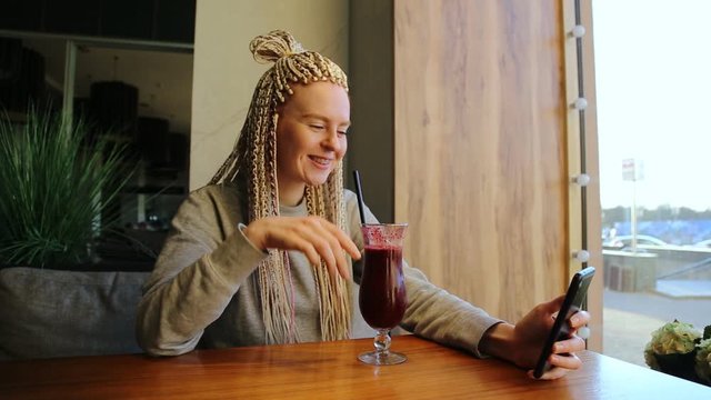 Cheerful woman blogger with kanekalons drinks berry smoothie and use phone.