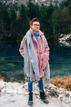 Cheerful woman in coat and denim trousers stands near the beautiful lake and smiles