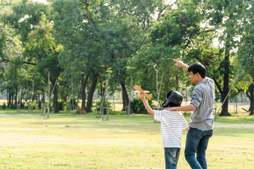Happy Asian father playing paper plane model with son at park, boy wearing pilot helmet, holding airplane toy in the air, Dad looking proud and support to his kid dream goal, family insurance concept