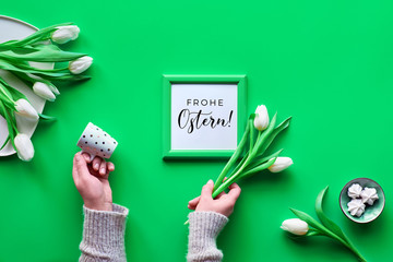 Hands with coffee cup and white Spring tulips, bunch of flowers on plate and marshmallow sweets. Neo mint green background, top view, frame with text "Frohe Ostern" that means Happy Easter in German.