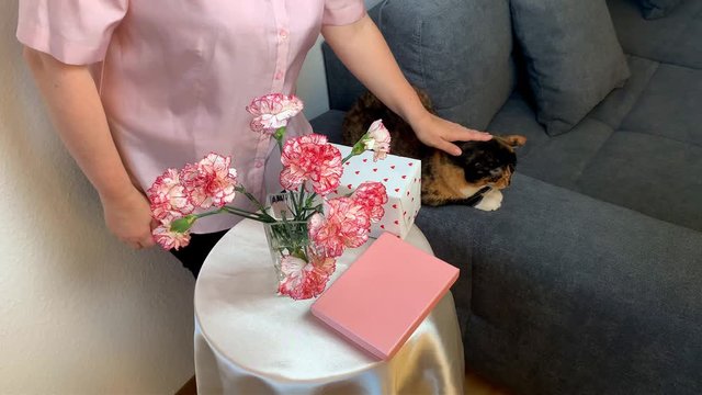 woman stroking a cat, a bouquet of pink flowers of carnations, smiling, gifts in boxes lie on a round table, cozy home concept