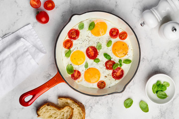 Fried eggs with tomatoes and basil in a red pan for breakfast on a marble table. Top view