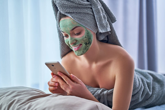Happy woman with green clay mask in bath towel and with smartphone is resting and relaxing during spa day at home