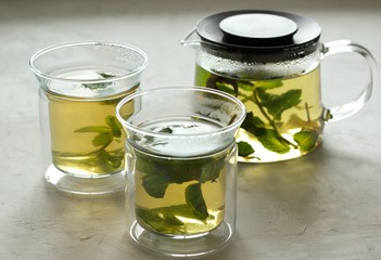 Two glass cups and a glass teapot with green tea with mint on a light gray table