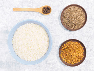 Indian long grain Basmati rice in blue ceramic bowl, allspice, cumin (jeera), fenugreek on white concrete background