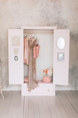wooden pink wardrobe for women’s clothes, open doors, decor, mirror, bag, hangers, silk dresses in a bright room, concept, flowers delicate pastel colors