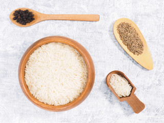 Indian long grain Basmati rice in brown wooden bowl, cumin (jeera) and black pepper in spoon on white concrete background