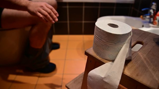 Close-up image of the toilet paper.In the blurred background you can recognize the bare legs of a Caucasian man as he defecates.The hands drum the fingers.The right hand tries to grab the toilet paper