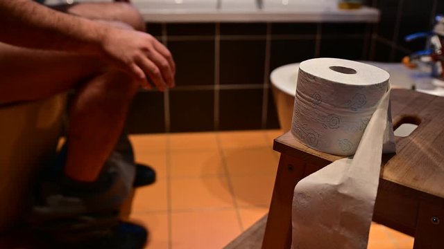 Close-up image of the toilet paper.In the blurred background you can recognize the bare legs of a Caucasian man as he defecates.The hands drum the fingers.The right hand tries to grab the toilet paper
