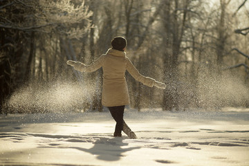 Young woman in white coat and black hat walking in sunny snowy forest at sunrise time in winter, frosty sunny winter