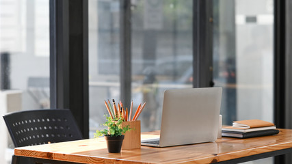 Photo of computer laptop, potted plant, pencil holder and books putting together on modern wooden table in the comfortable office with wall glass as background. Comfortable working space concept.