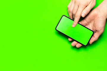 Male hands hold a modern black smartphone with green blank screen on neon green background flat lay top view. Modern technology, phone, gadget in hands, touch screen, template for your design. Mockup