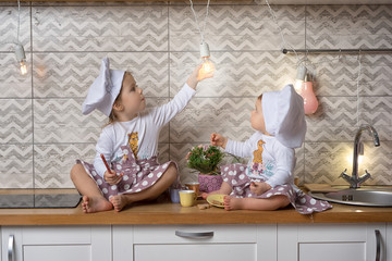 Two sisters one-year-old and three-years-old girls in a cook hats sit on the kitchen table and...