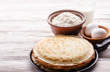Stack of French crepes in frying pan on wooden kitchen table with milk eggs and flour aside