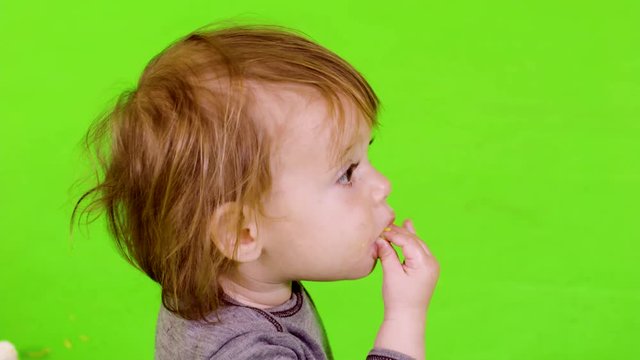 Adorable little toddler laughing while she playing and eating with her hands, looking at the camera, close-up, over green screen.