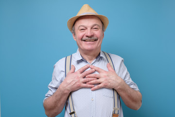Cheerful senior man smiles happily, keeps hands on chest. Pleased male glad to recieve compliment...