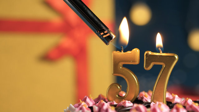 Birthday cake number 57 golden candles burning by lighter, background gift yellow box tied up with red ribbon. Close-up