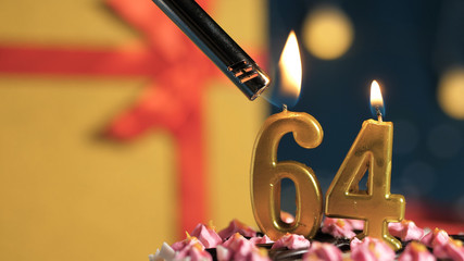 Birthday cake number 64 golden candles burning by lighter, background gift yellow box tied up with...