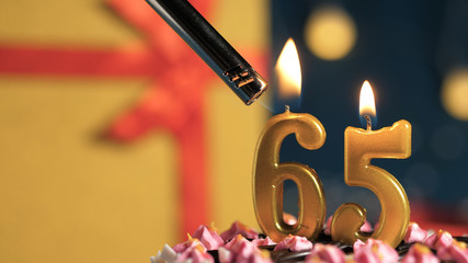 Birthday cake number 65 golden candles burning by lighter, background gift yellow box tied up with...