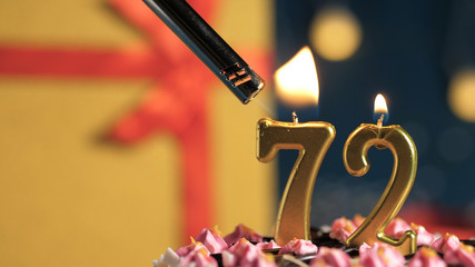 Birthday cake number 72 golden candles burning by lighter, background gift yellow box tied up with...