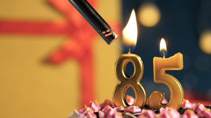 Birthday cake number 85 golden candles burning by lighter, background gift yellow box tied up with...