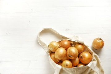 A lot of fresh organic yellow onions in a reusable textiles bag on a white wooden background.Zero waste shopping, plastic free, ecological concept.Close-up, top view, copy space.