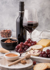 Bottle and glass of red wine with selection of various cheese in wooden box and grapes on light table background. Blue Stilton, Red Leicester and Brie Cheese with Cheddar and nuts with honey.