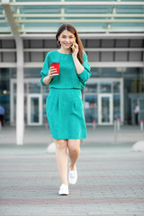 Young adult brunette speaks smatfonu and cross the road. Hot coffee in a plastic glass. Green dress. The concept of business, work, lifestyle and technology.