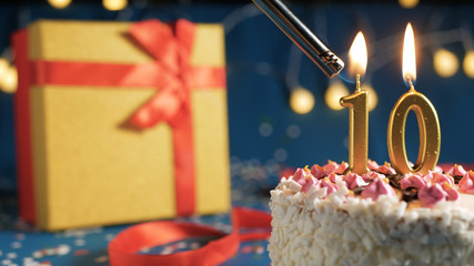 White birthday cake number 10 golden candles burning by lighter, blue background with lights and...