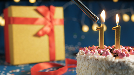 White birthday cake number 11 golden candles burning by lighter, blue background with lights and gift yellow box tied up with red ribbon. Close-up