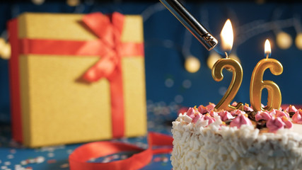 White birthday cake number 26 golden candles burning by lighter, blue background with lights and gift yellow box tied up with red ribbon. Close-up