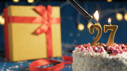 White birthday cake number 27 golden candles burning by lighter, blue background with lights and gift yellow box tied up with red ribbon. Close-up