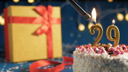White birthday cake number 29 golden candles burning by lighter, blue background with lights and...