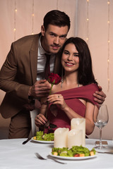 handsome man hugging happy girlfriend holding red rose while looking at camera