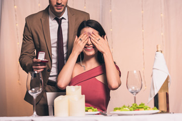 elegant man holding wedding ring near happy girlfriend sitting at served table and covering eyes with hands