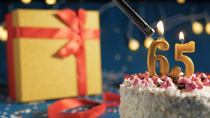 White birthday cake number 65 golden candles burning by lighter, blue background with lights and...