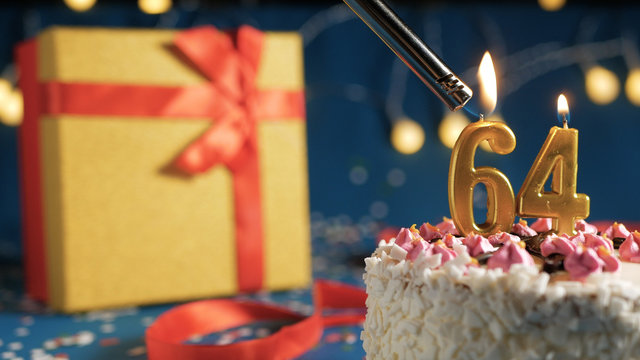White birthday cake number 64 golden candles burning by lighter, blue background with lights and gift yellow box tied up with red ribbon. Close-up