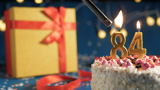 White birthday cake number 84 golden candles burning by lighter, blue background with lights and gift yellow box tied up with red ribbon. Close-up