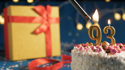 White birthday cake number 93 golden candles burning by lighter, blue background with lights and gift yellow box tied up with red ribbon. Close-up