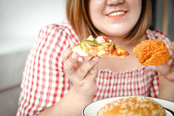 An Asian fat woman is eating pizza and fried chicken on the sofa in her house. The concept of consuming food that causes disease is not good for health