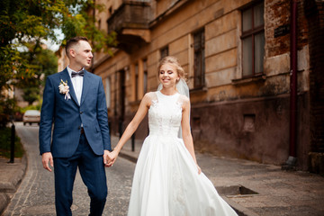 Wedding photo shooting. Bride and bridegroom walking in the city. Married couple embracing and looking at each other. Holding bouquet. Outdoor, full body